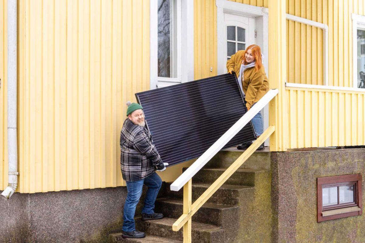 Aurinkosähkö tulee taloon! Mies ja nainen kantavat aurinkopaneelia talon portaita.